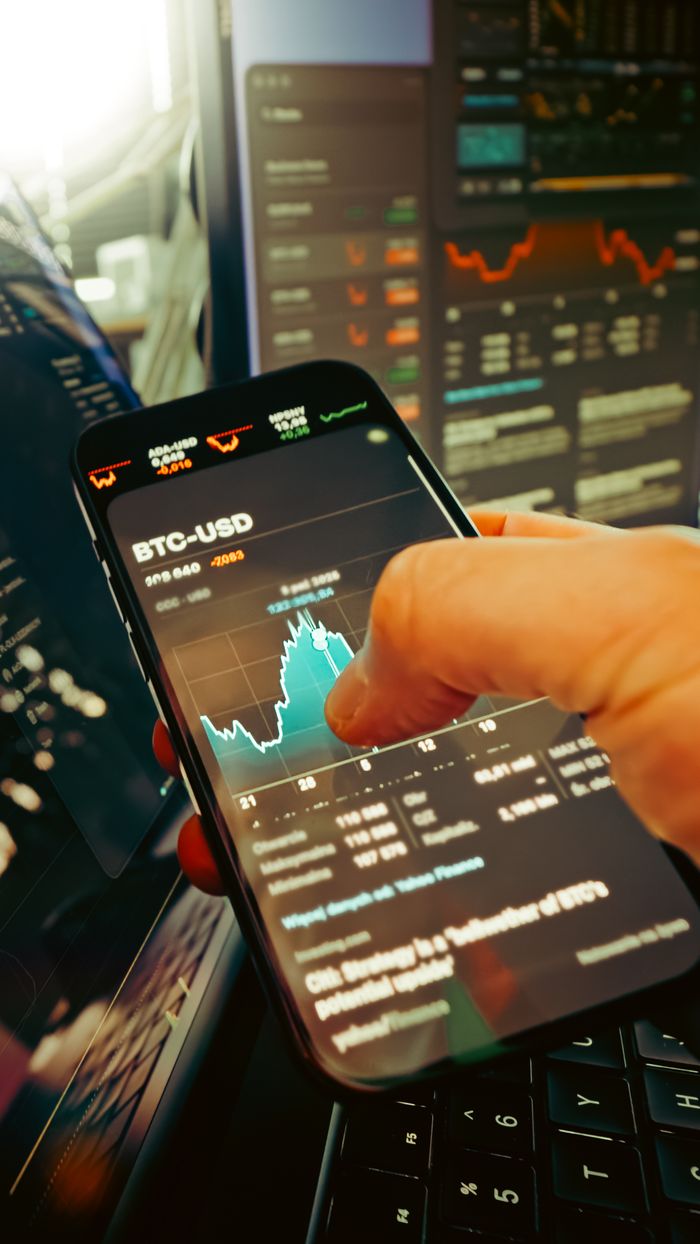 about-04 A hand holds a smartphone displaying BTC-USD chart amidst digital trading screens, highlighting crypto finance.